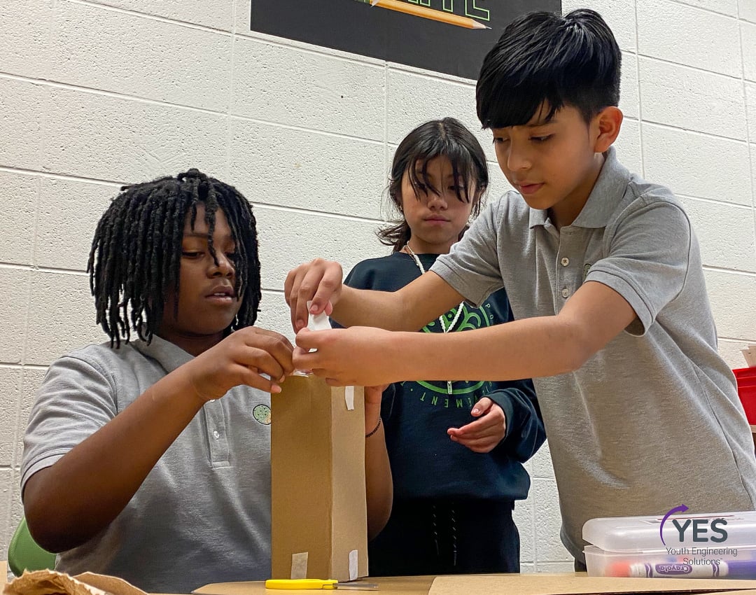 Three students build a tall rectangular box during a STEM activity.